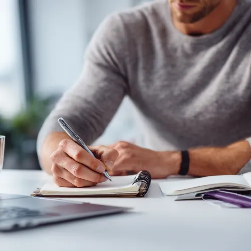 نمای نزدیک از یک مرد جوان در حال نوشتن در دفتر یادداشت روی میز کار؛ تصویری از فرایند تدوین، تمرکز و خلق محتوا در تحریریه وب‌سایت باشگاه مدیران و کارآفرینان مثلث.