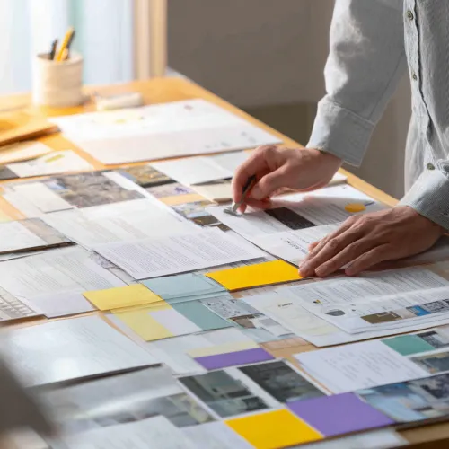 نمایی از دستان یک فرد در حال مرور و چیدمان اسناد، یادداشت‌ها و برگه‌های مختلف روی میز؛ نمادی از تحلیل، سازمان‌دهی محتوا و برنامه‌ریزی در تحریریه وب‌سایت باشگاه مدیران و کارآفرینان مثلث.