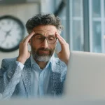 مدیر میانسال با عینک و موهای جوگندمی پشت لپ‌تاپ، دست‌ها روی شقیقه‌ها و نشانه‌های فشار کاری؛ تصویر مرتبط با سلامت روان مدیران و هزینه پنهان بی‌توجهی به تعادل کاری.