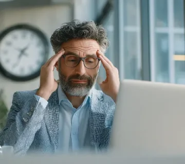 مدیر میانسال با عینک و موهای جوگندمی پشت لپ‌تاپ، دست‌ها روی شقیقه‌ها و نشانه‌های فشار کاری؛ تصویر مرتبط با سلامت روان مدیران و هزینه پنهان بی‌توجهی به تعادل کاری.