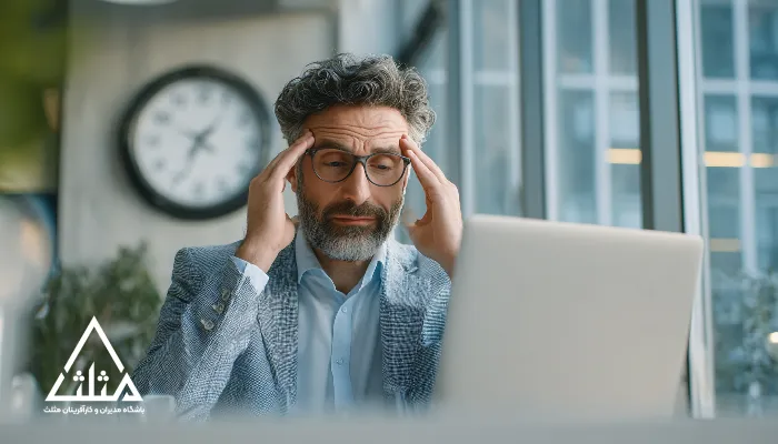 مدیر میانسال با عینک و موهای جوگندمی پشت لپ‌تاپ، دست‌ها روی شقیقه‌ها و نشانه‌های فشار کاری؛ تصویر مرتبط با سلامت روان مدیران و هزینه پنهان بی‌توجهی به تعادل کاری.